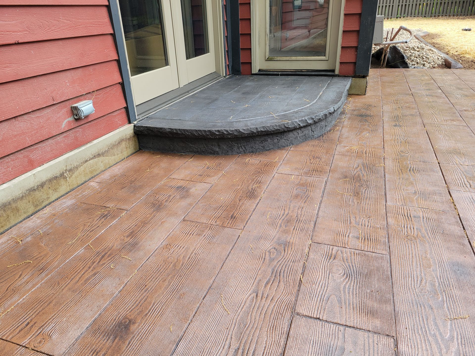 A semi-circular, dark grey stone step sits outside a door on a patio with brown, wood-grain stamped concrete planks.