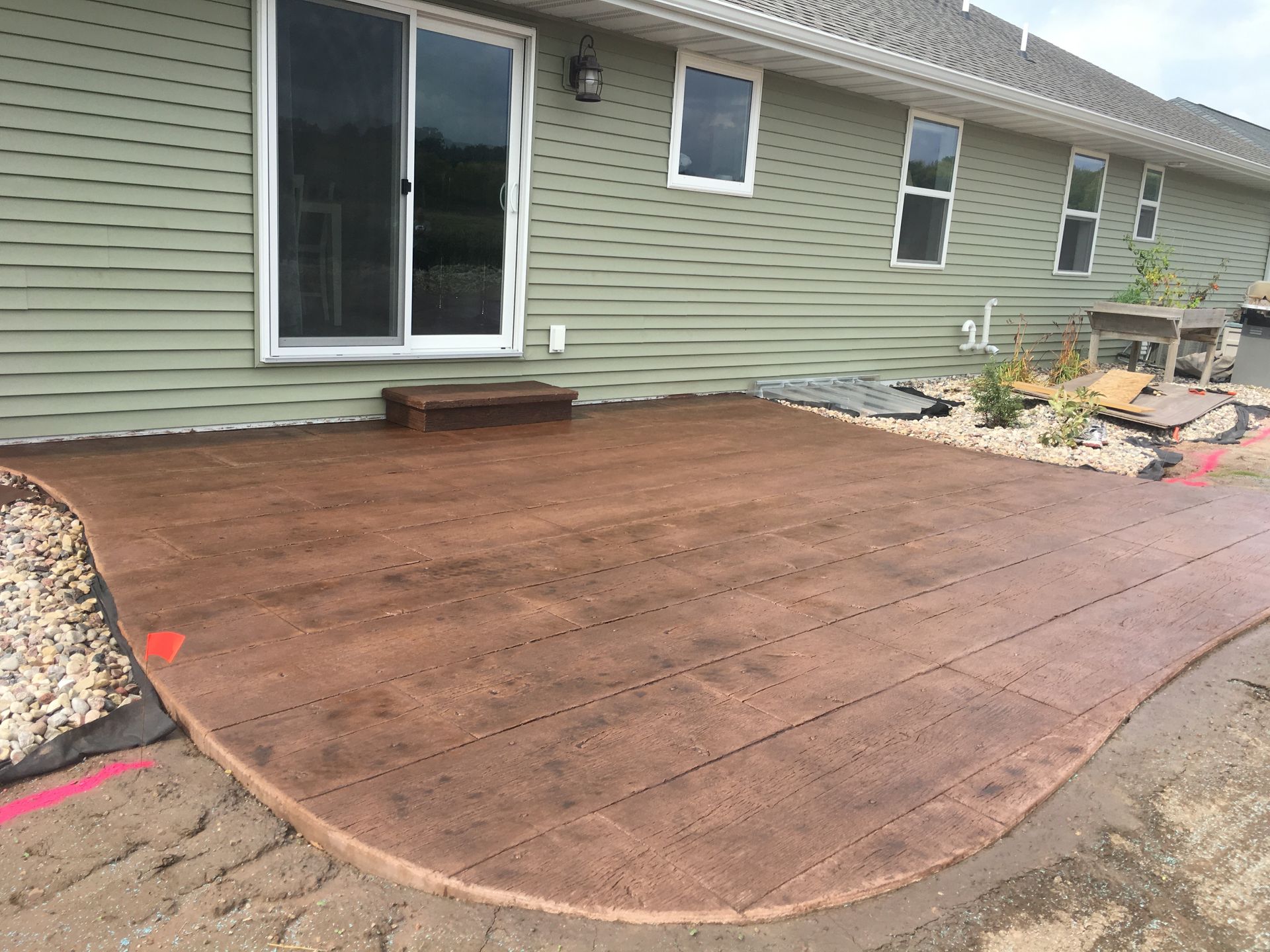 A new brown, stamped concrete patio with a rounded edge installed against the green siding of a house.