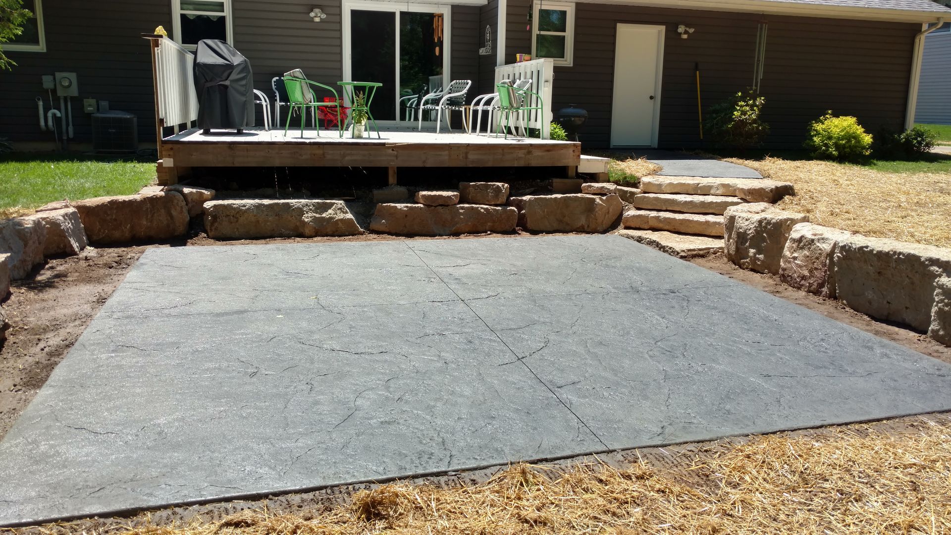 A patio area with a rectangular concrete slab, stone retaining walls, and a wooden deck with furniture in the background.