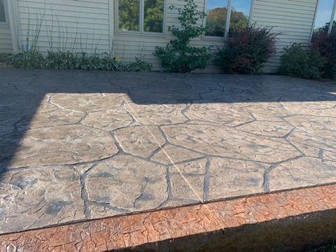 A close-up of a stamped concrete patio with a flagstone pattern, featuring a textured red brick border at the base.