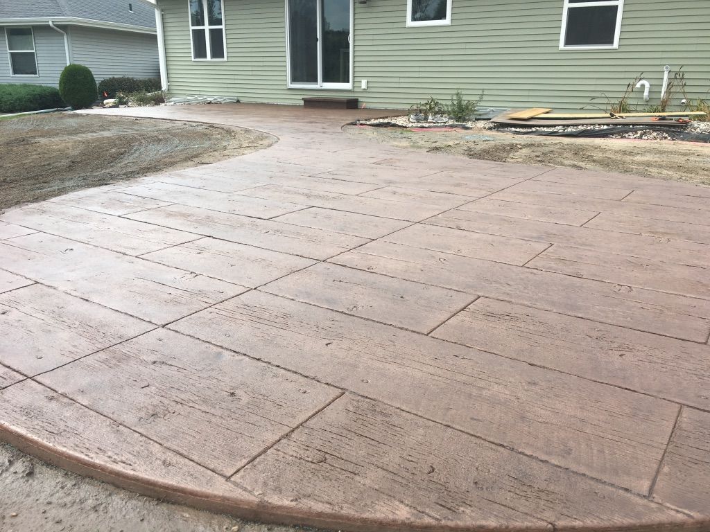 A tan, stamped concrete patio with a grid pattern, extending from the back sliding doors of a house with green siding.