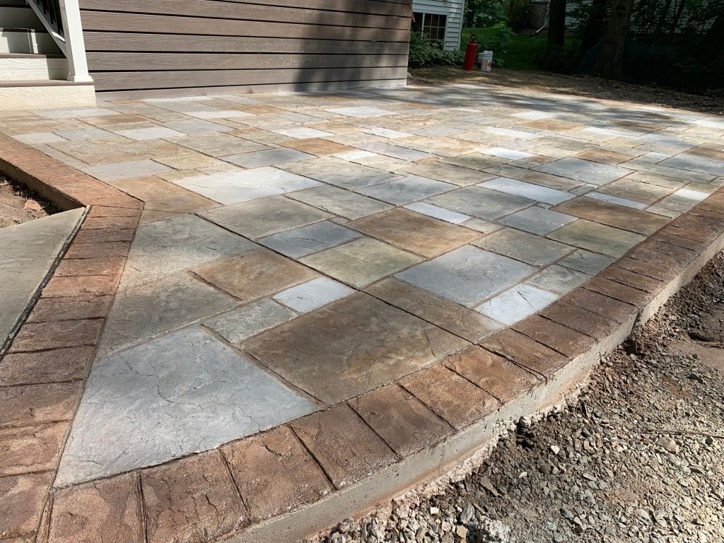 A ground-level view of a newly installed patio featuring stone pavers with a decorative brick border, beside a house deck.