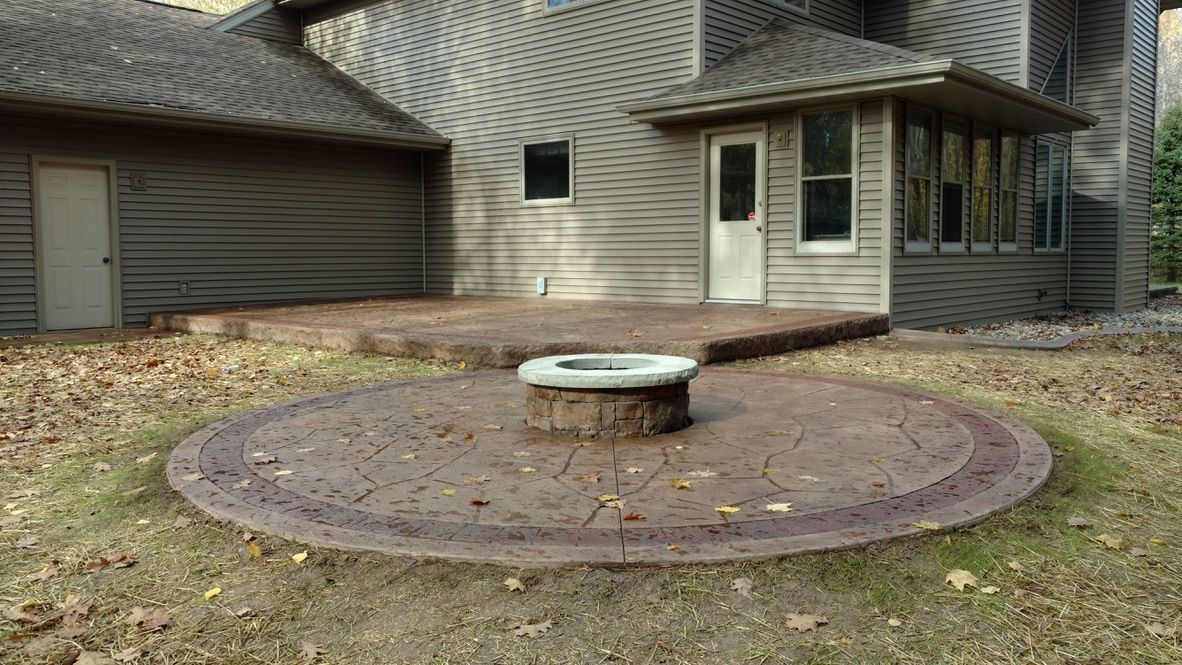 A round stamped concrete patio with a central stone fire pit in front of a gray house exterior during autumn.
