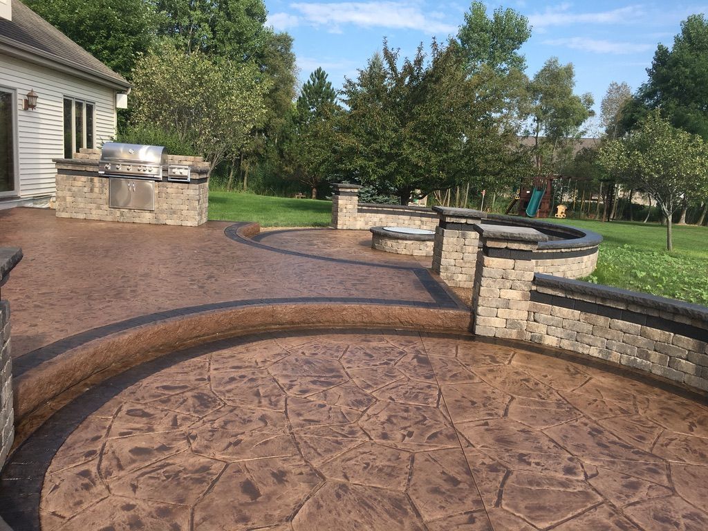 A multi-level stamped concrete patio with a stone grill island, curved retaining walls, and surrounding lawn.