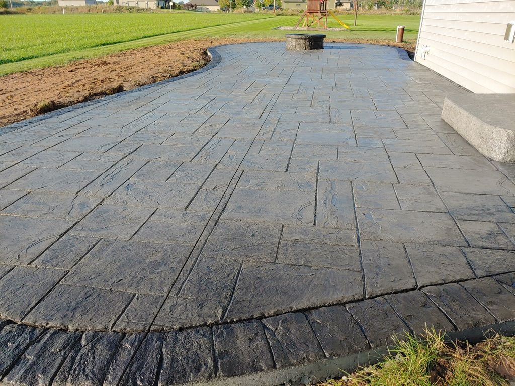 A gray stamped concrete patio with a dark border, fire pit, and adjacent grassy yard next to a house exterior.