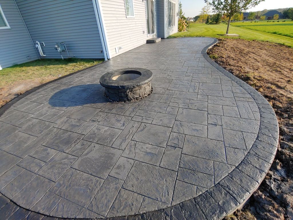 A grey, stamped concrete patio with a circular stone fire pit, bordered by a dark paver edge next to a house.