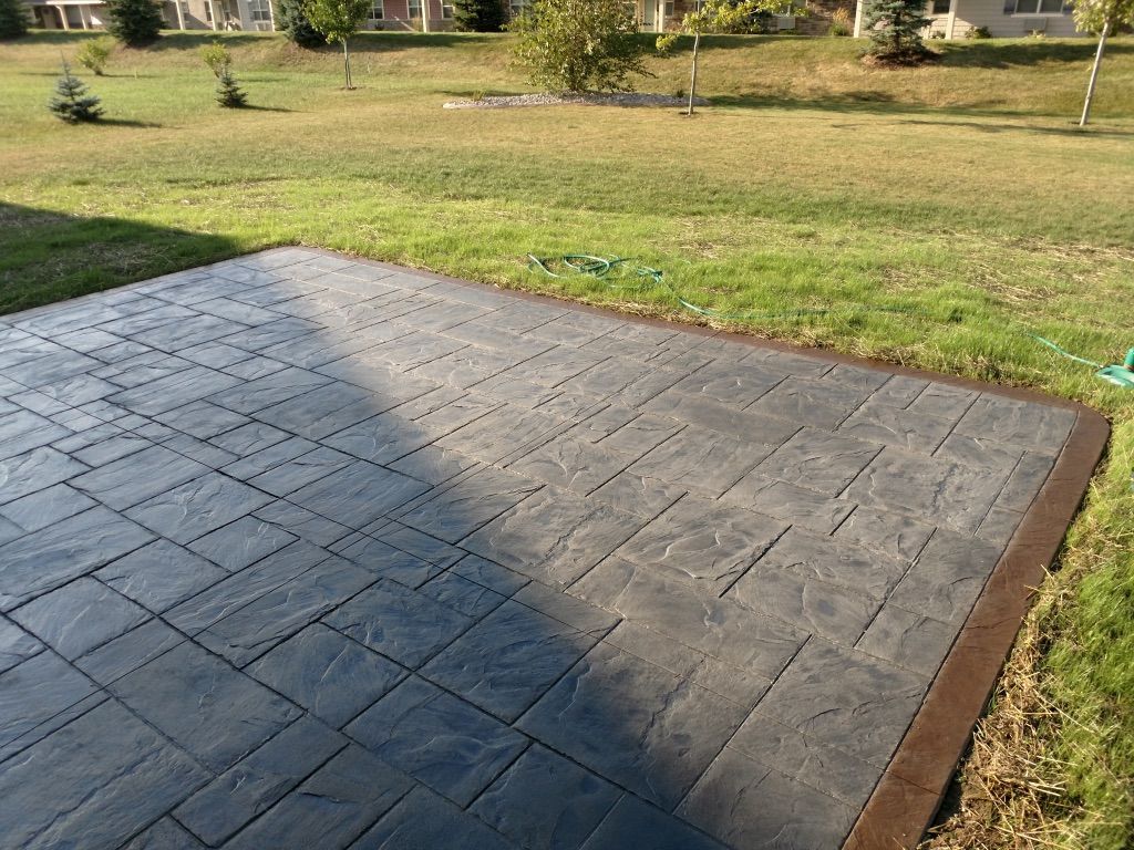 A gray stamped concrete patio with a dark border, set against a green lawn and trees in a residential backyard.