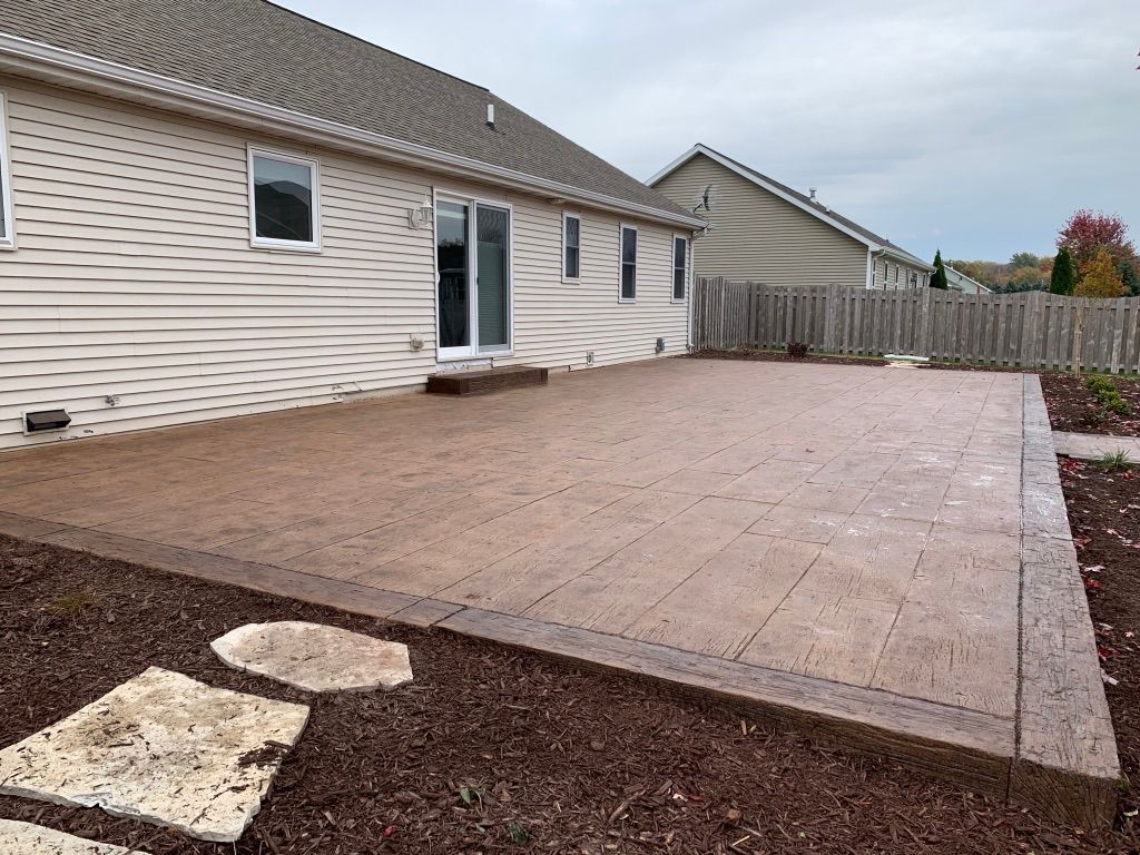 A wide tan-colored stamped concrete patio featuring a darker border, adjacent to a light-sided house with a sliding door.