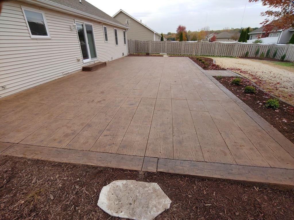 A stamped concrete patio with a wood-plank pattern adjacent to a beige house, with mulch and a stone stepping stone.