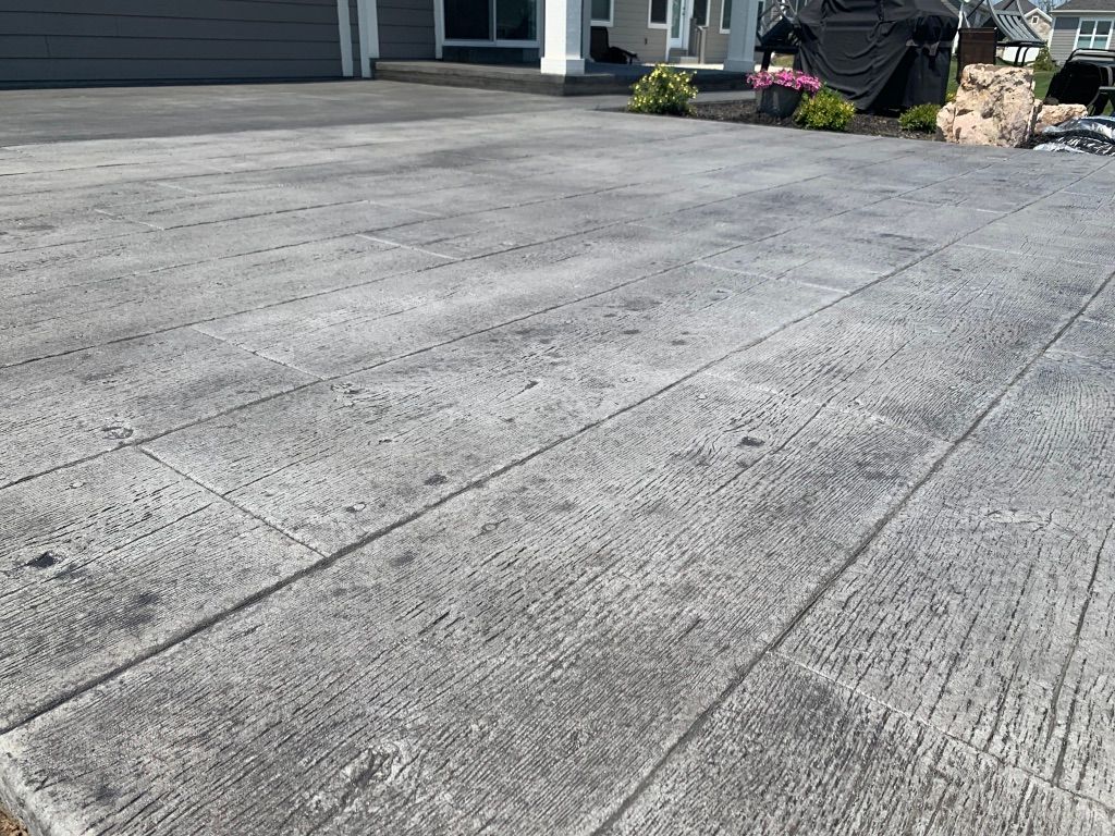 A textured gray concrete patio with stamped linear patterns, situated near a house with landscaping and a grill.