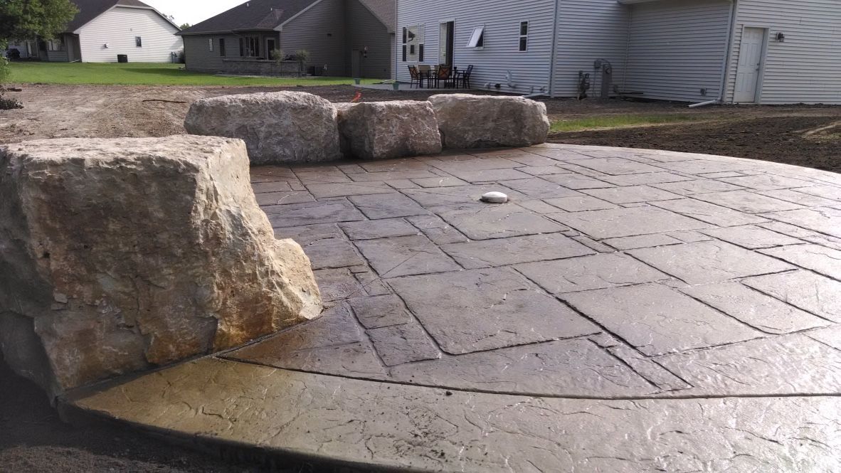 A curved, stamped concrete patio with large, natural stone blocks bordering the back edge in a residential backyard.