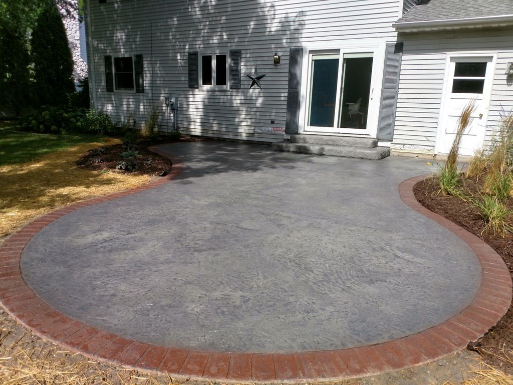A gray stamped concrete patio with a red brick border, situated in a backyard beside a white house with a sliding door.