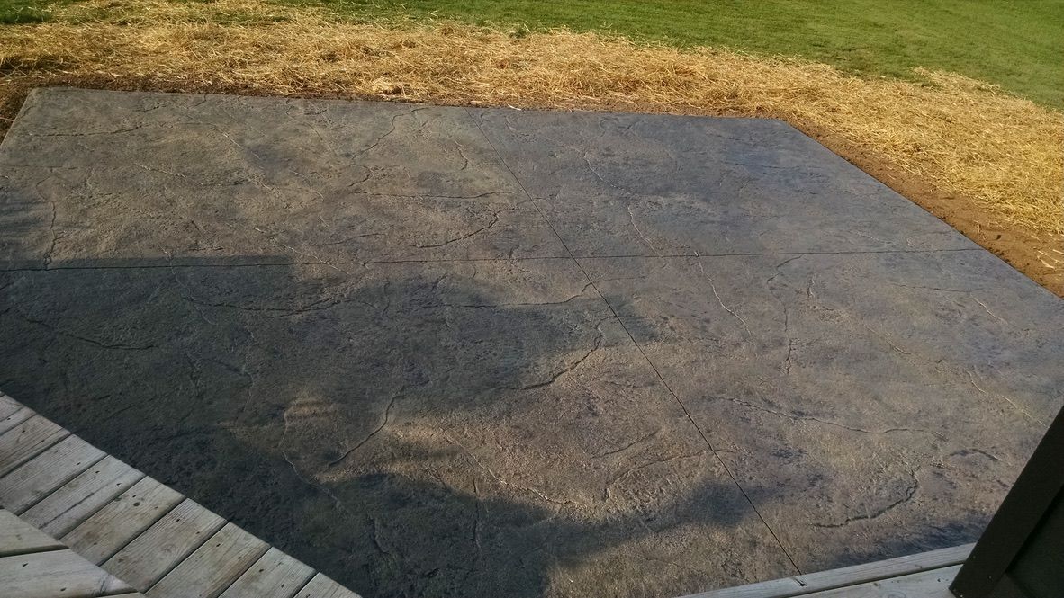 A dark, rectangular patio area made of textured stamped concrete next to a wooden deck and grass.