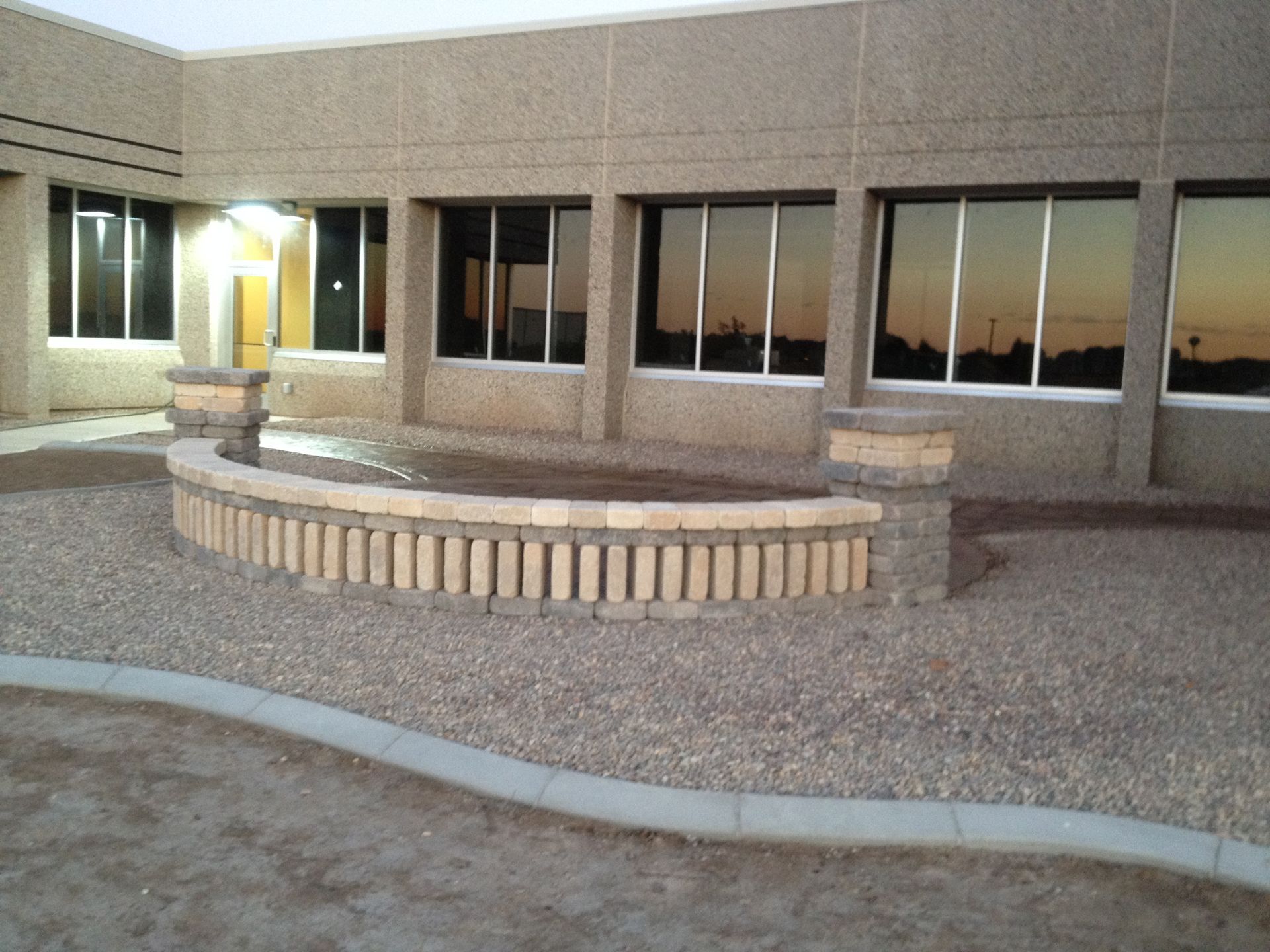 A curved stone seating wall sits in a gravel courtyard in front of a modern commercial building at sunset.