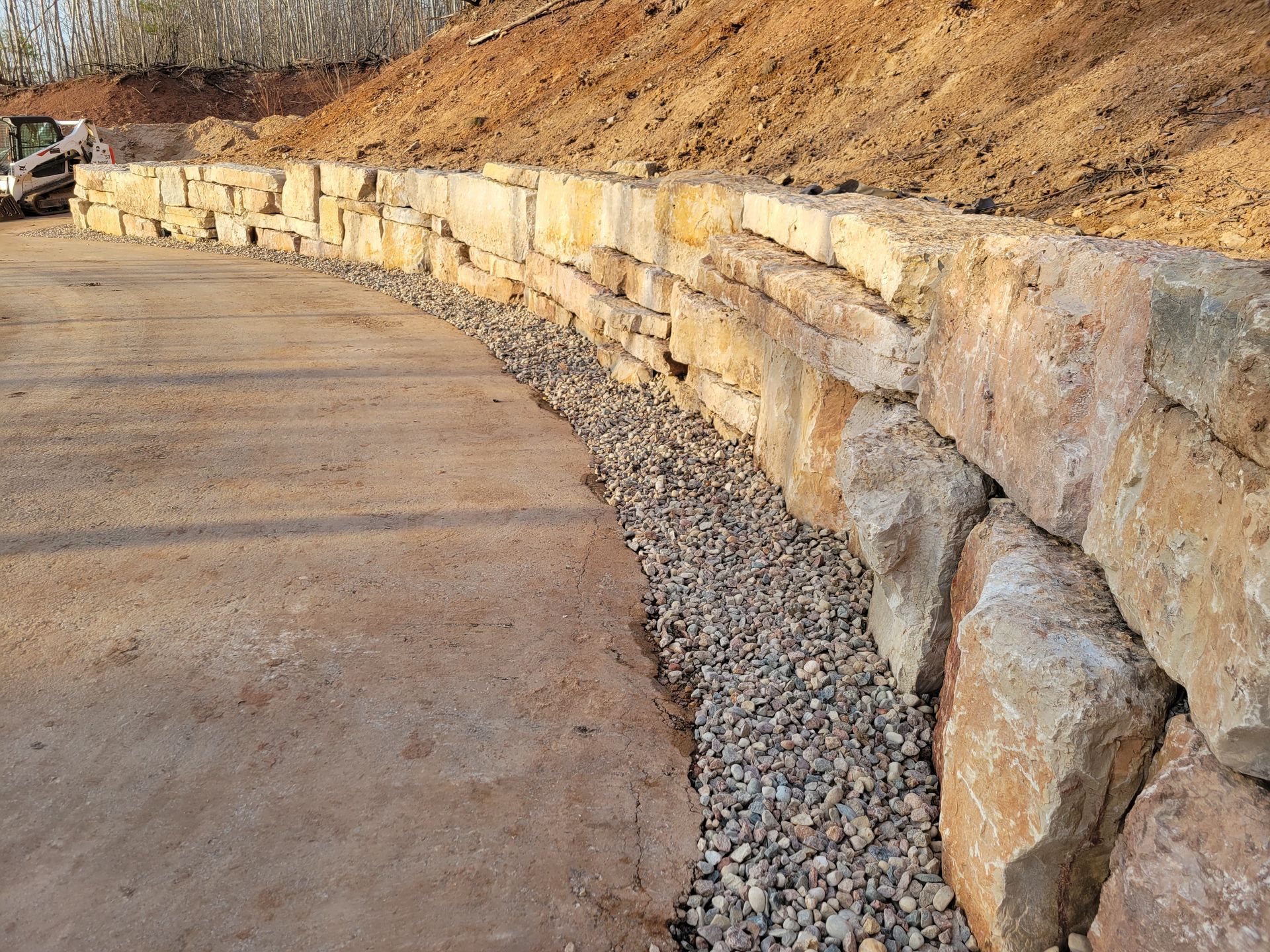 A curved retaining wall made of large, stacked sandstone blocks, bordering a gravel drainage strip and a dirt path.