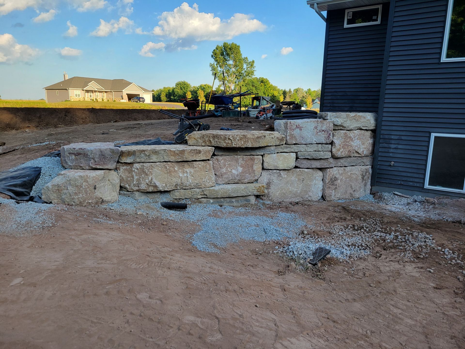 A stone block retaining wall under construction next to a dark-sided house with a dirt yard and a distant home.