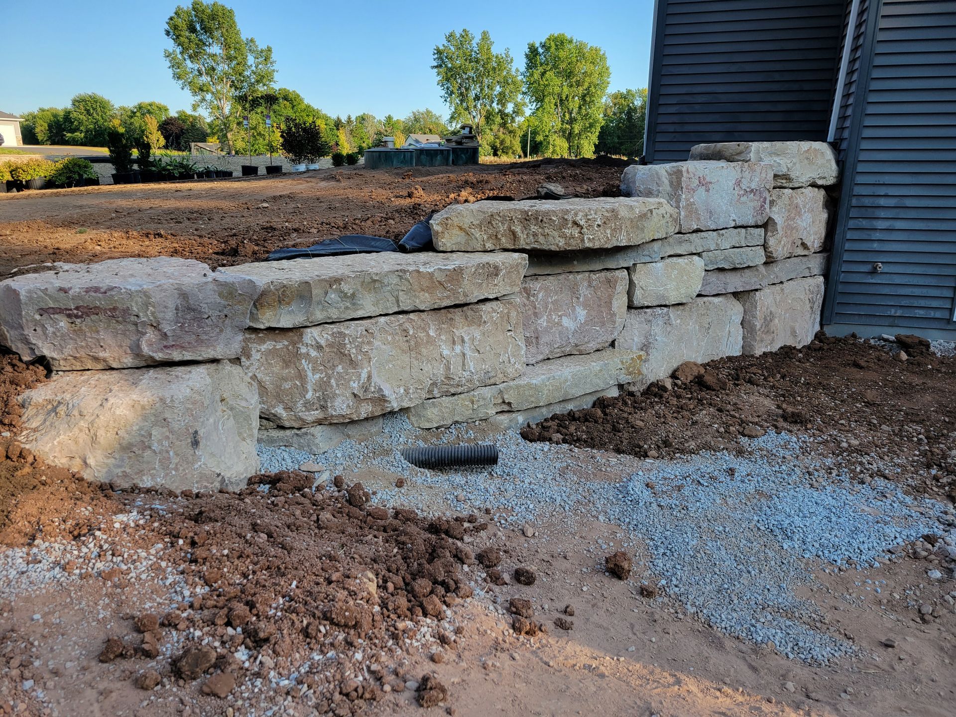 A newly constructed stone retaining wall made of large, light-colored blocks sits beside a dark house in a dirt yard.