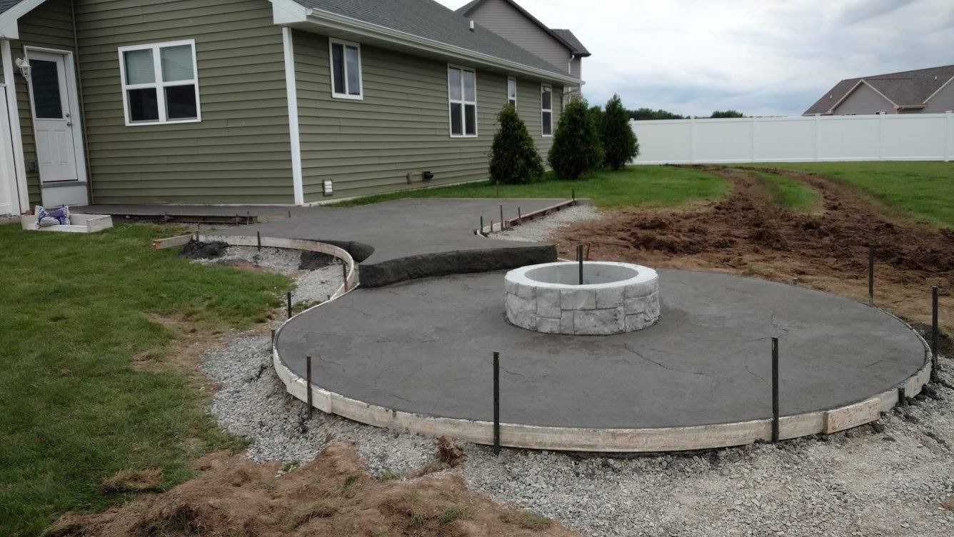 A newly poured circular concrete patio with a stone fire pit sits in a backyard next to an olive-green house.