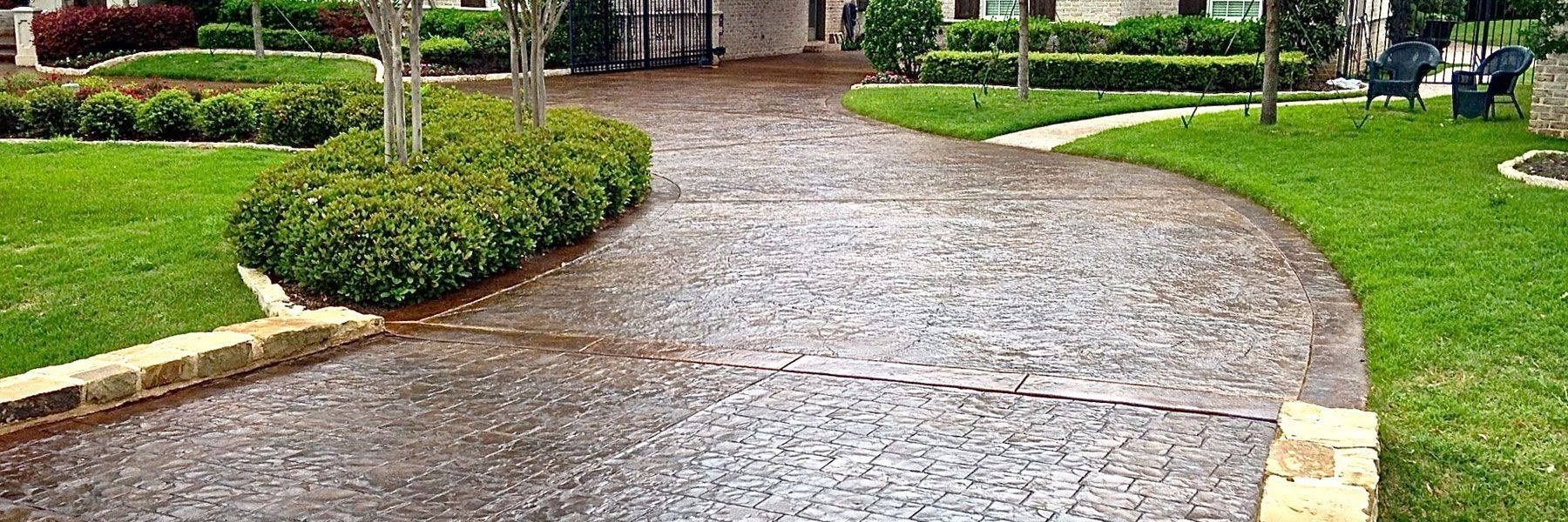 A paved driveway with a textured surface leads toward a house, framed by manicured green lawns and sculpted shrubbery.