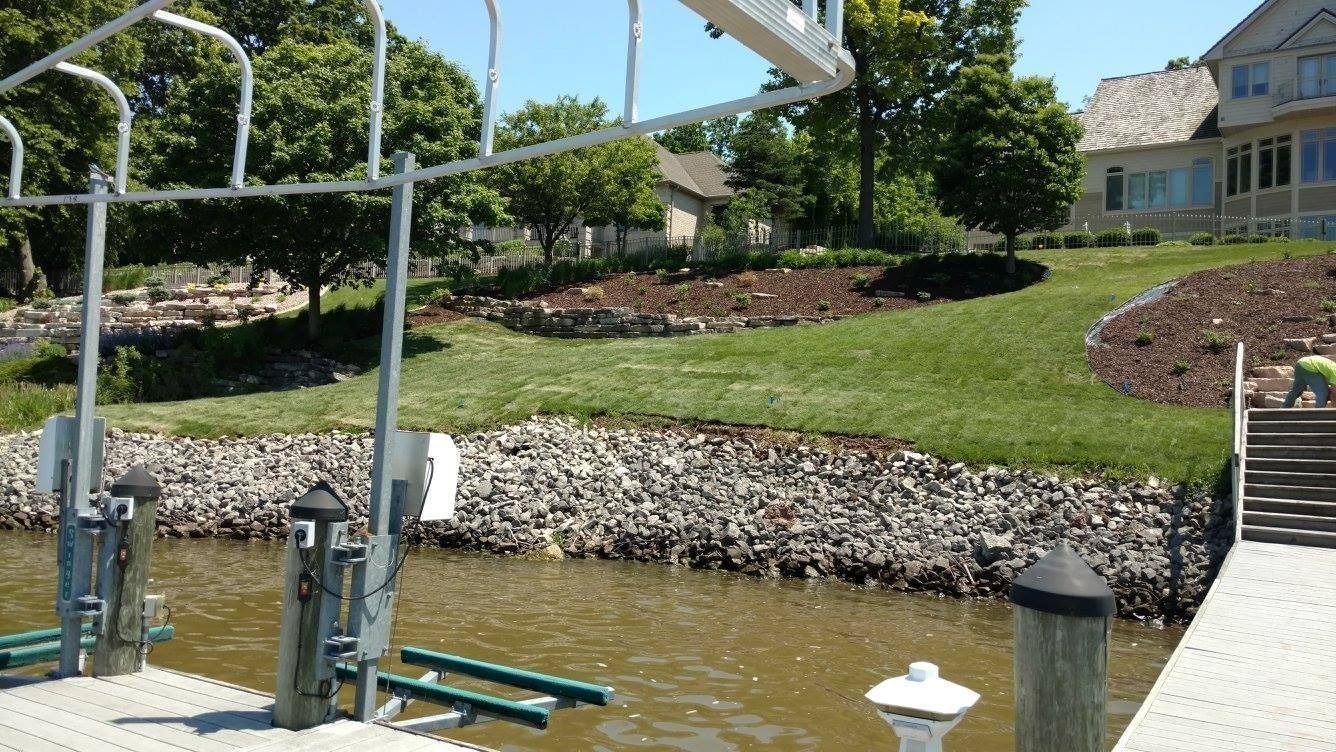 A boat lift and dock extend into a lake, with a stone seawall, grassy lawn, landscaping, and a house in the background.