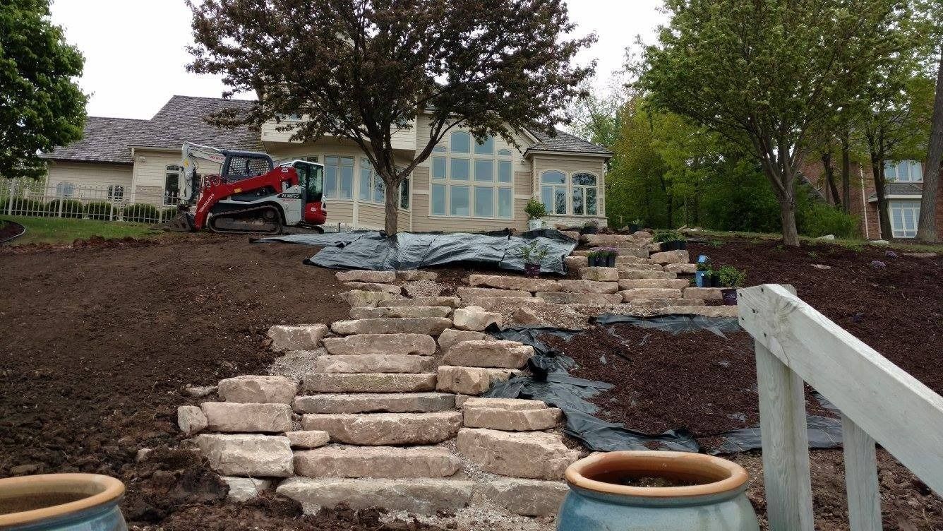 A stone staircase under construction leads up a sloped yard toward a house with a red skid steer parked nearby.