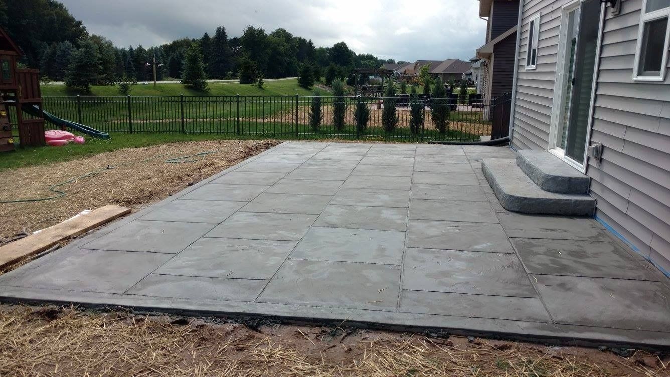 A grey, rectangular concrete patio recently installed outside a house with siding and a short set of concrete steps.