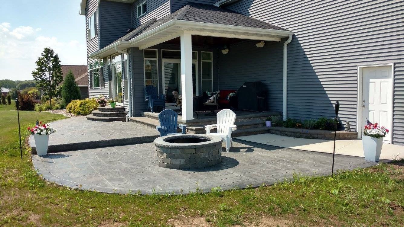 A gray house with a stone patio featuring a built-in fire pit, outdoor furniture, and two white potted flower planters.