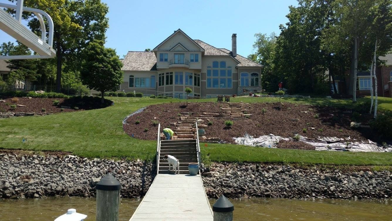 A house overlooks a lake, viewed from a wooden dock where a white dog stands at the base of stone stairs.