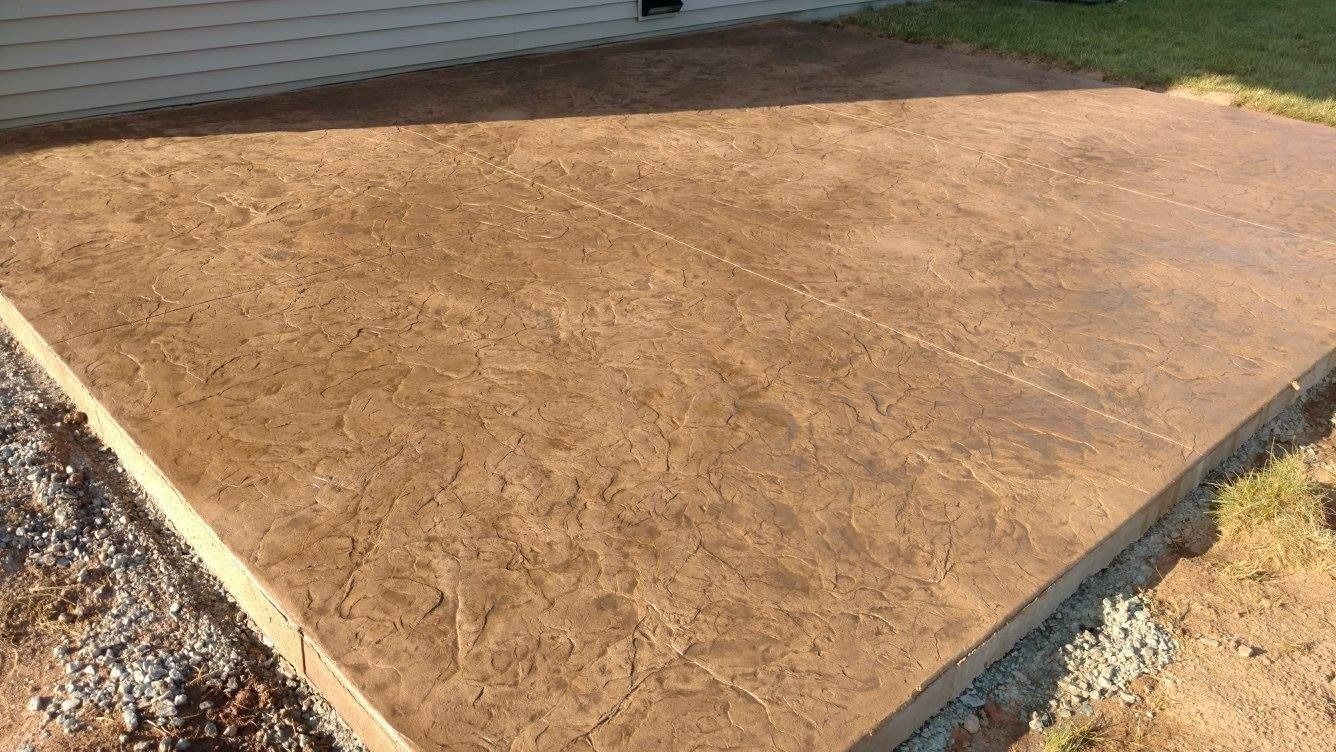 A newly poured brown stamped concrete patio adjacent to a light-colored house wall, with dirt and gravel along the edges.