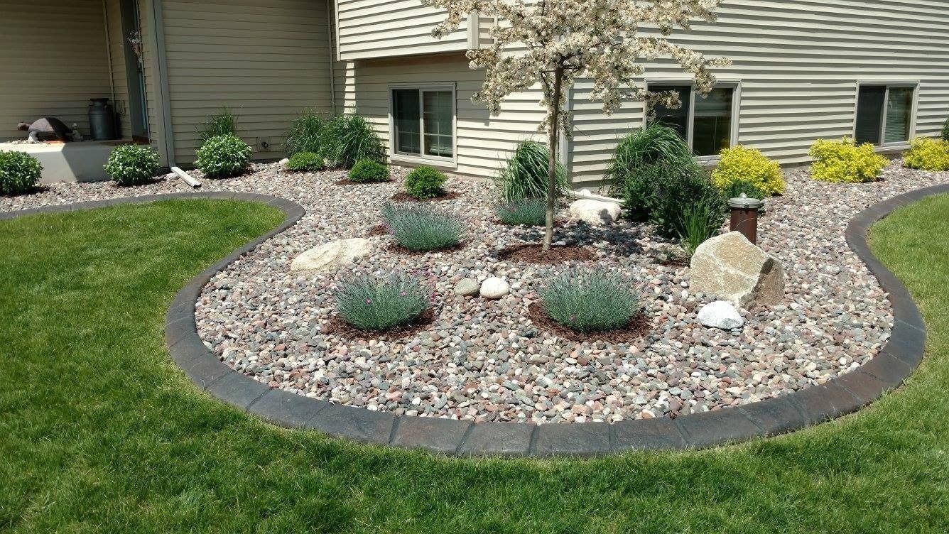 A landscaped garden bed filled with grey river rock, featuring small shrubs, a flowering tree, and stone accents.