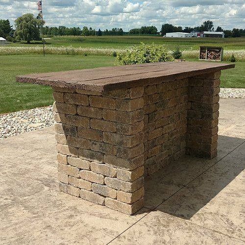 A tan, stone brick outdoor bar with a rustic wooden countertop, situated on a patio overlooking a green field.