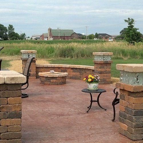 A brick-paved patio with a circular stone fire pit, seating, and a small metal table overlooking a grassy field.