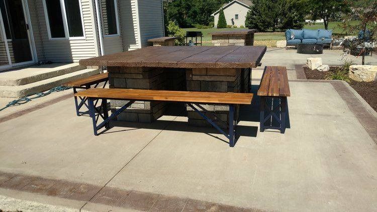 A large, square wooden patio table with stone bases and matching benches on a concrete surface outside a house.