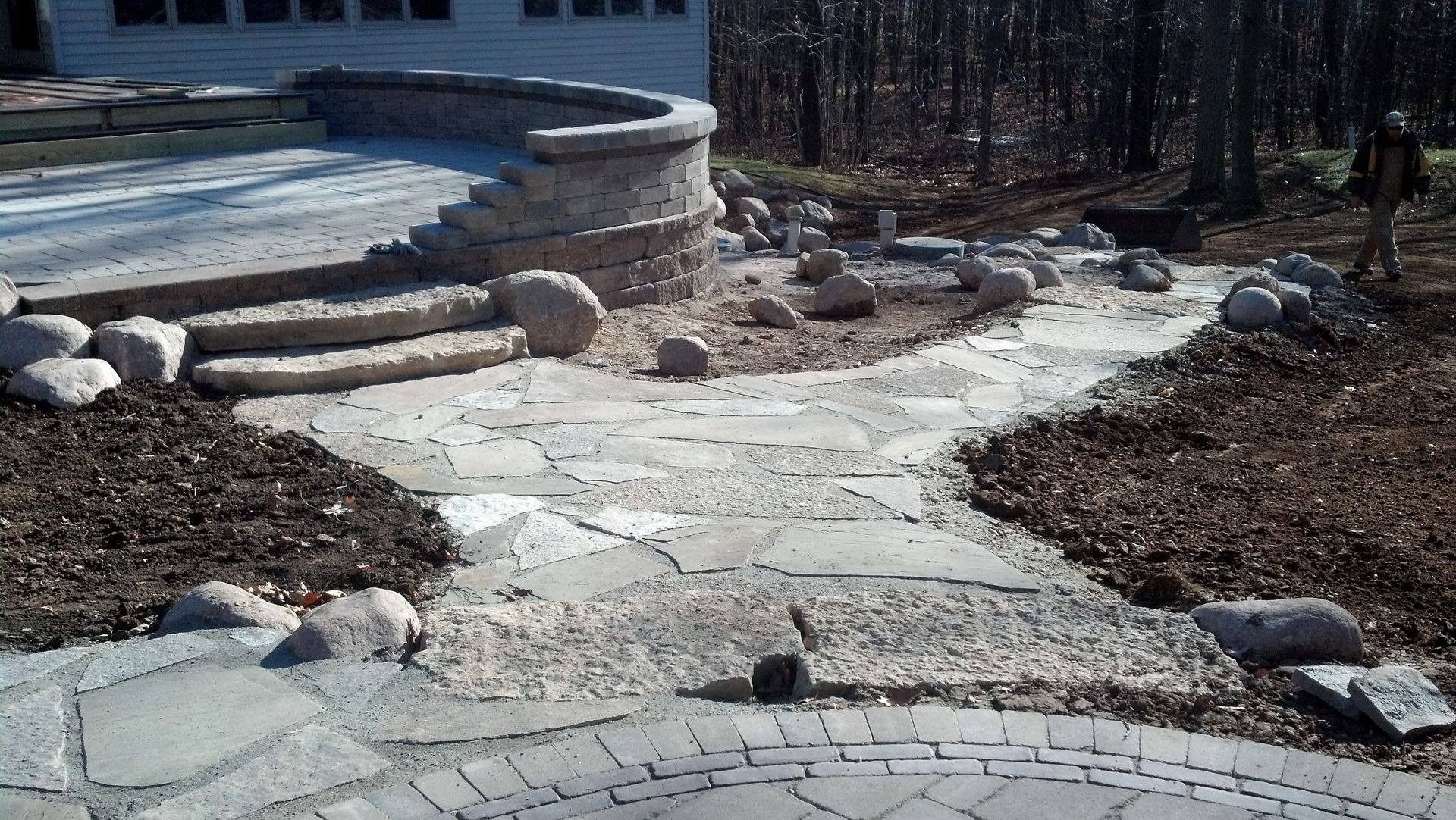 A stone patio and walkway lead to curved steps and a tiered retaining wall set against a wooded backdrop.