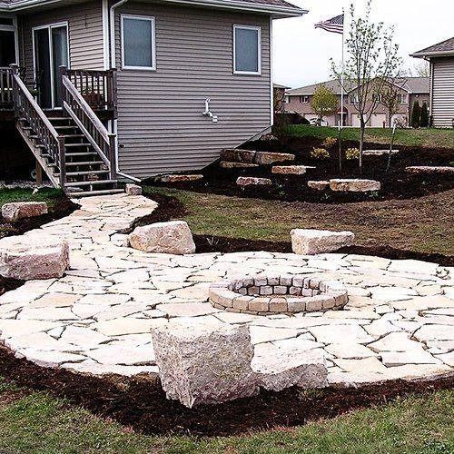 A stone flagstone patio with a round fire pit in a backyard next to a house with light gray siding and wooden stairs.