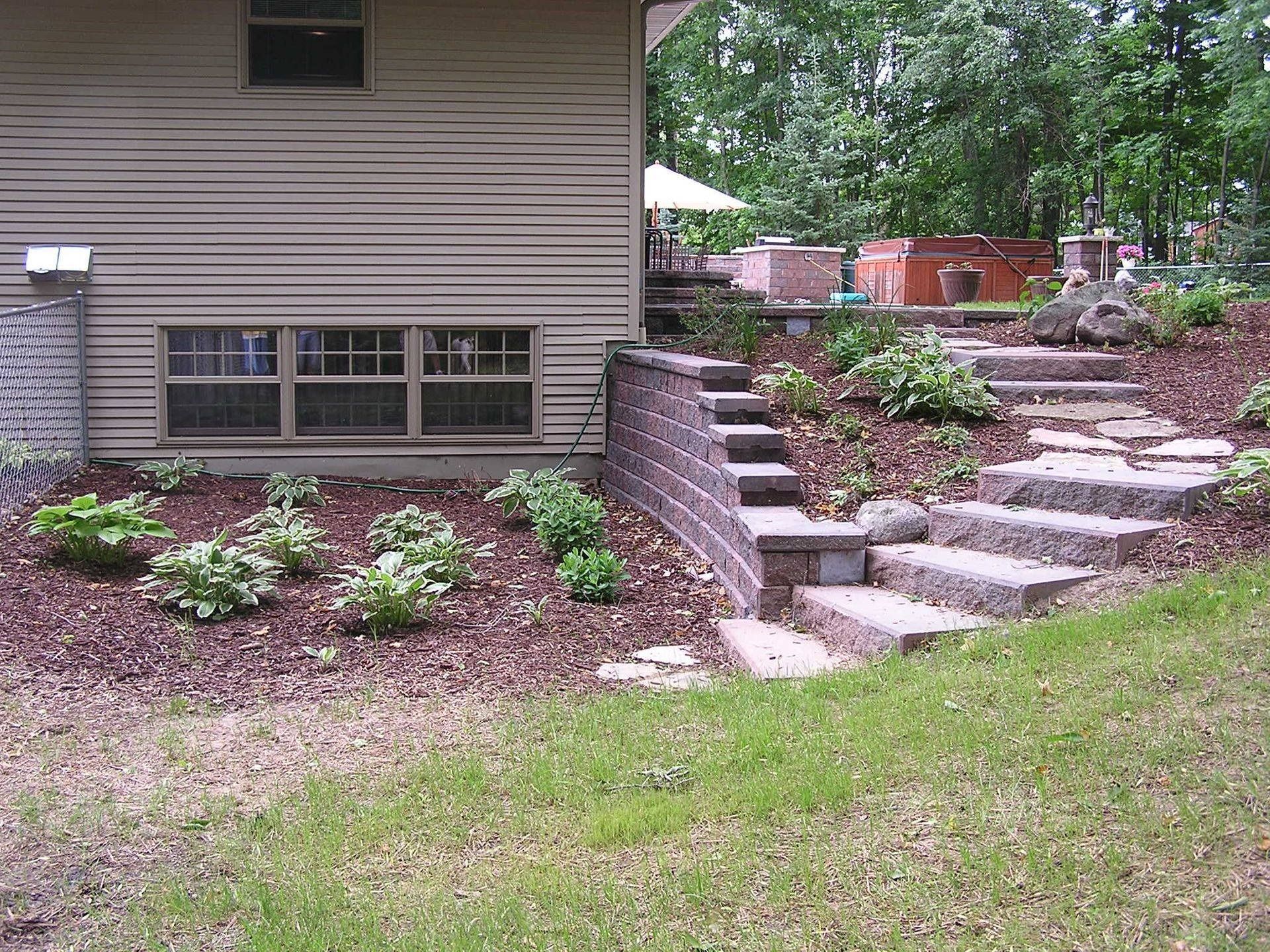 A tan house with a low-level window, next to a terraced stone retaining wall and steps leading to a backyard garden.
