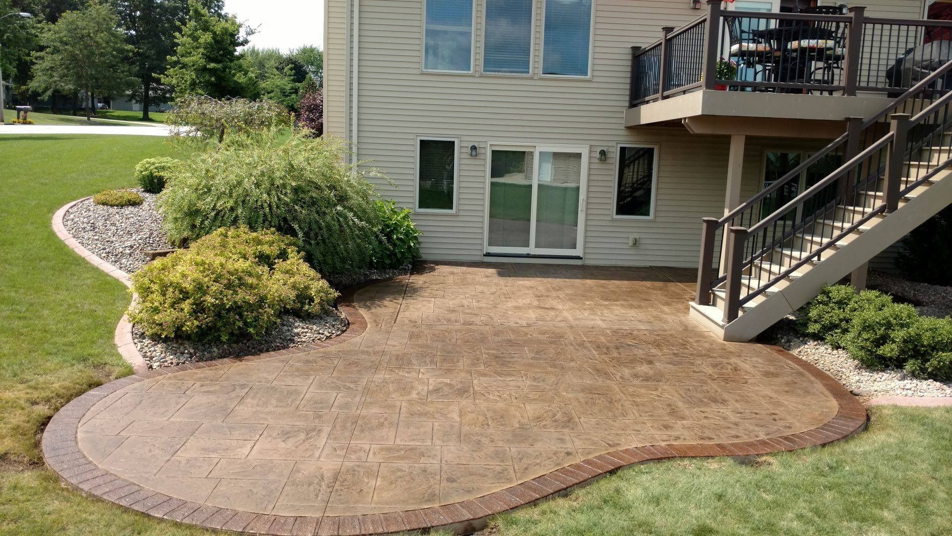 A brown-toned paver patio with a curved decorative border set against the back of a beige house with a wooden deck.