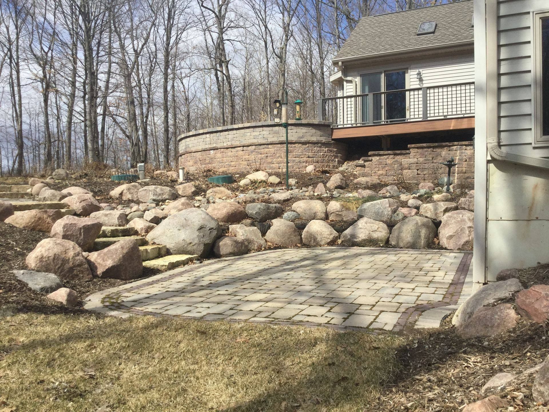 A stone patio with steps leading up to a tiered retaining wall and a house deck in a wooded yard.