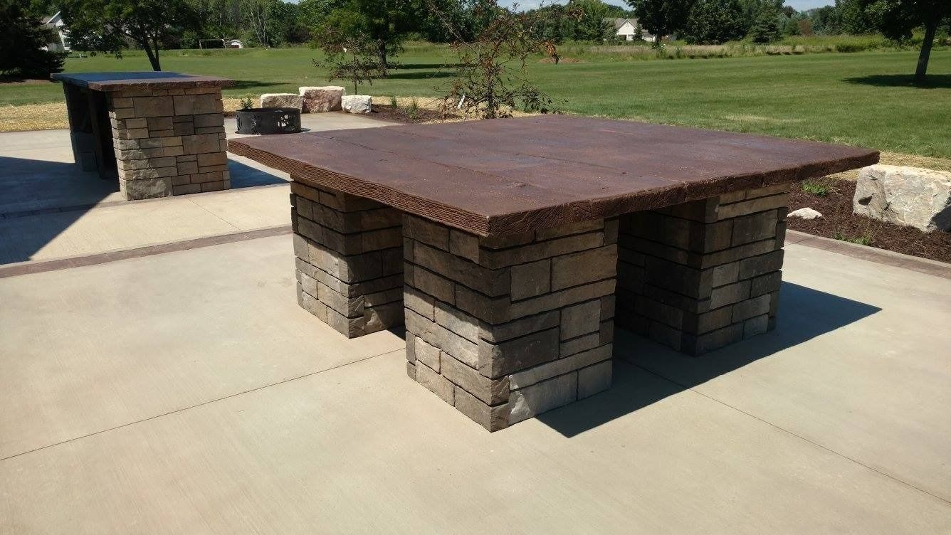 A square table with a dark stone top and a stacked-stone pedestal base on a concrete patio in a grassy yard.