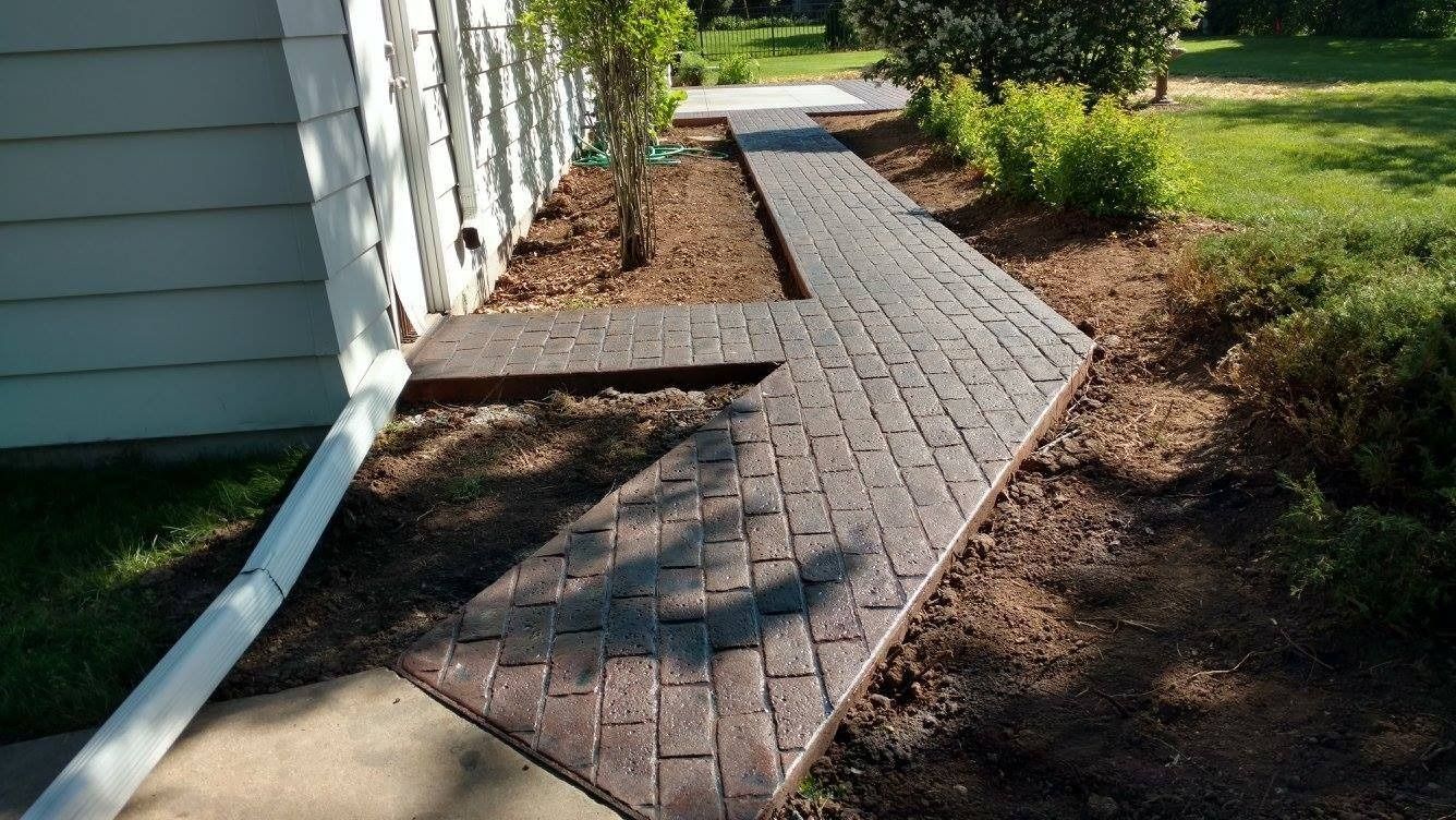 A newly installed dark-toned brick paver walkway runs alongside the side of a house next to a mulch bed.