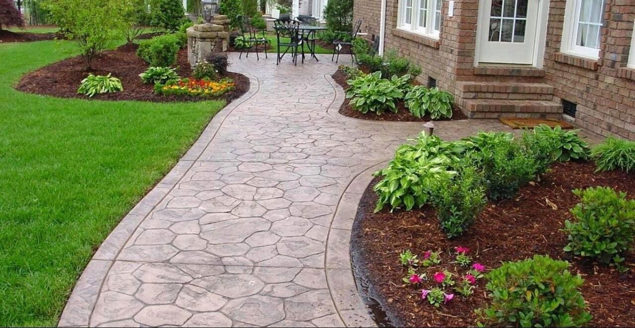 A stamped concrete walkway leads to a brick house entrance, flanked by green lawns, garden beds with mulch, and shrubs.