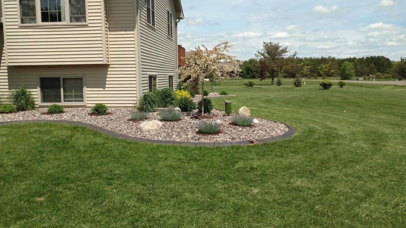 A beige house corner with a curved rock garden bed containing small shrubs, decorative stones, and a small tree.