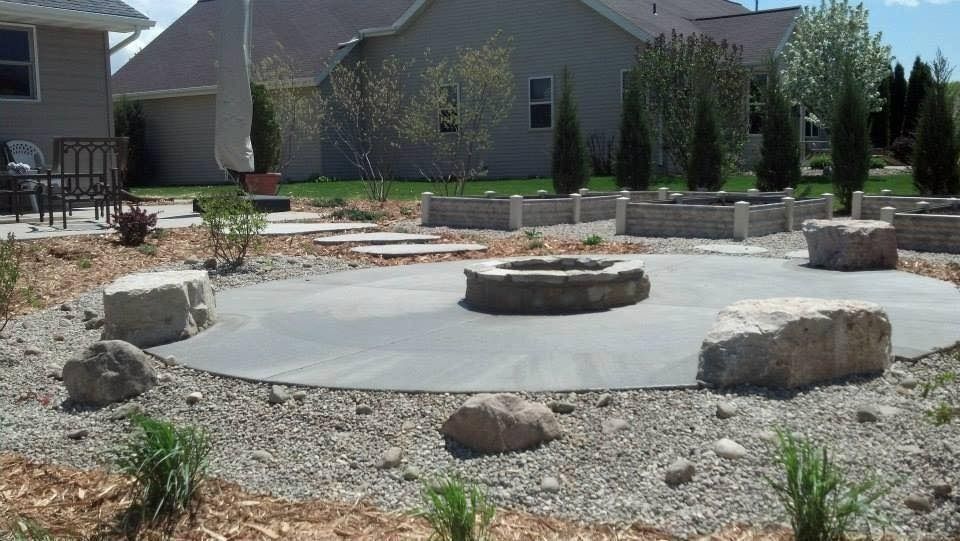 A backyard patio with a central stone fire pit, surrounded by gravel, decorative boulders, and raised garden beds.