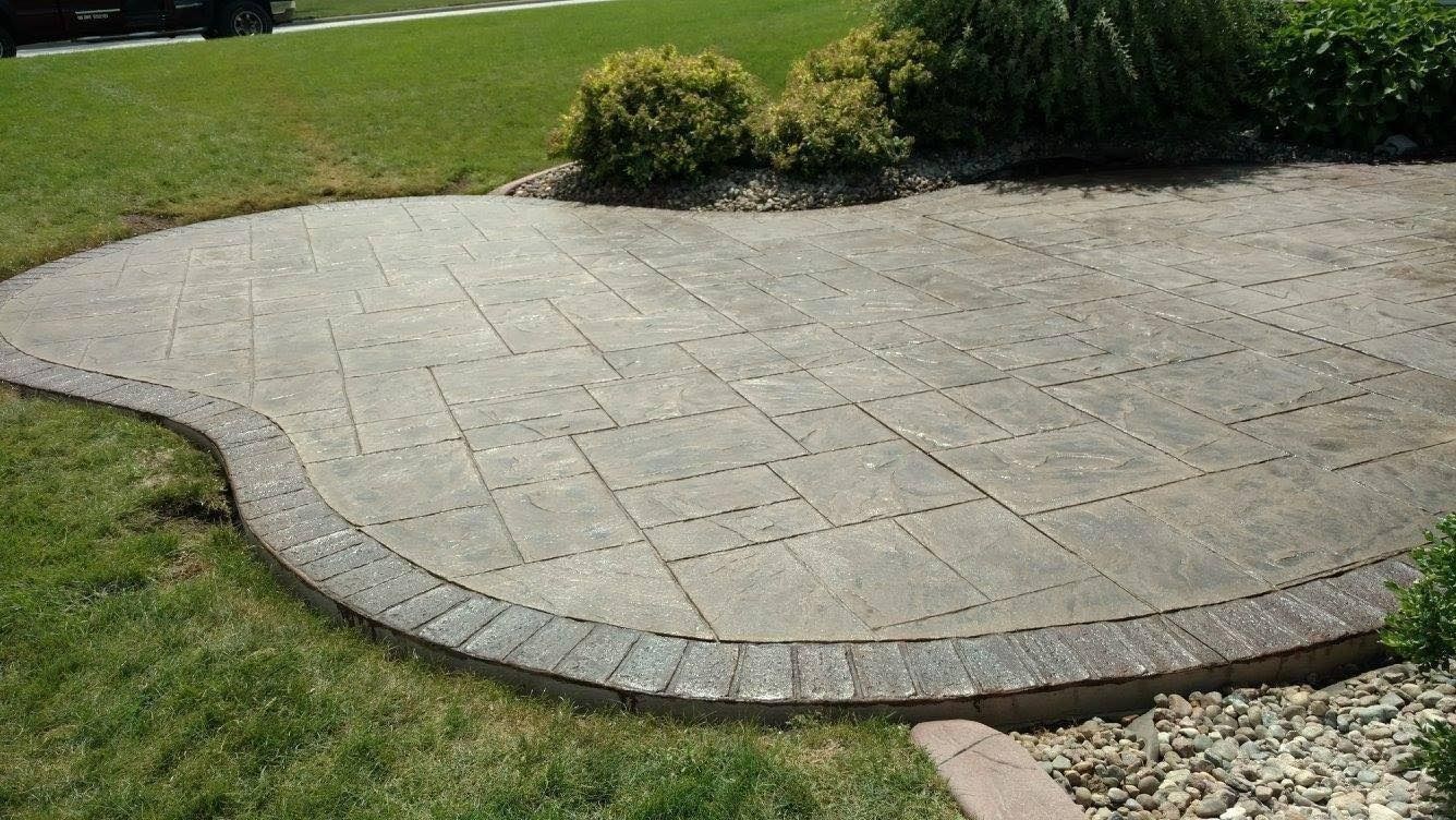 A curved stone patio with a textured, rectangular paver pattern and a darker border, set next to a grassy lawn and shrubs.