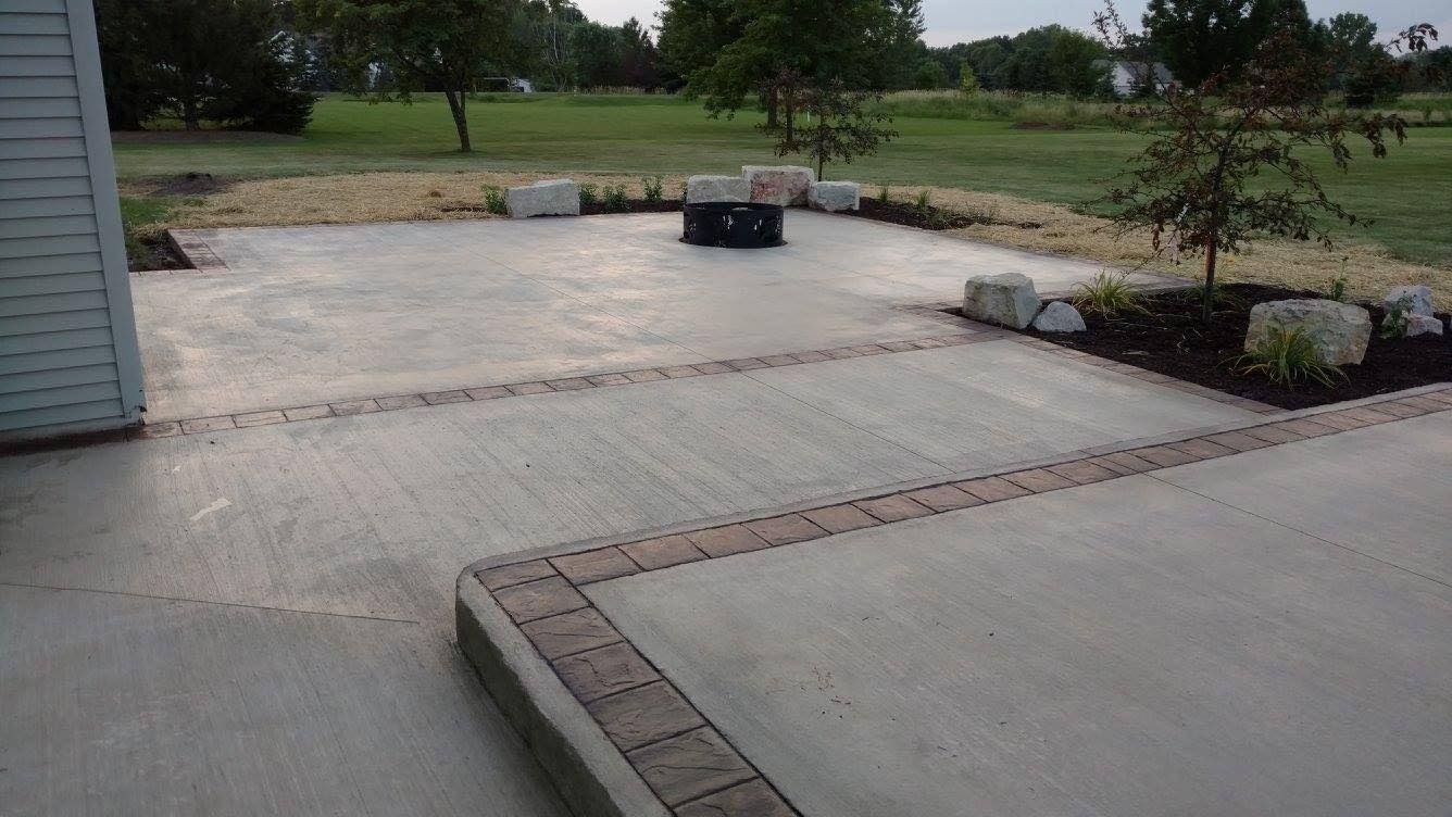 A concrete patio with a decorative brick border, a fire pit, and surrounding garden beds with large decorative rocks.