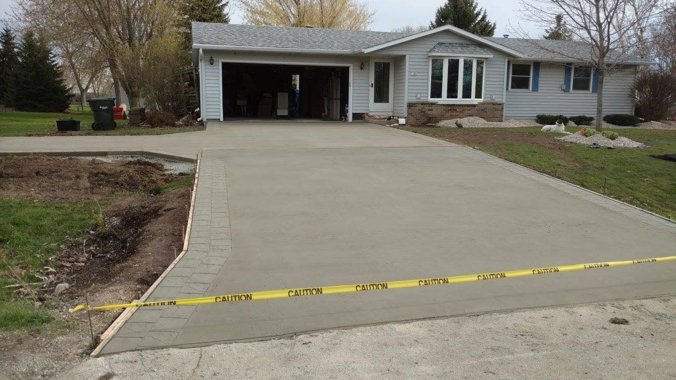 A newly poured concrete driveway leads to a suburban ranch-style house, sectioned off by yellow caution tape.