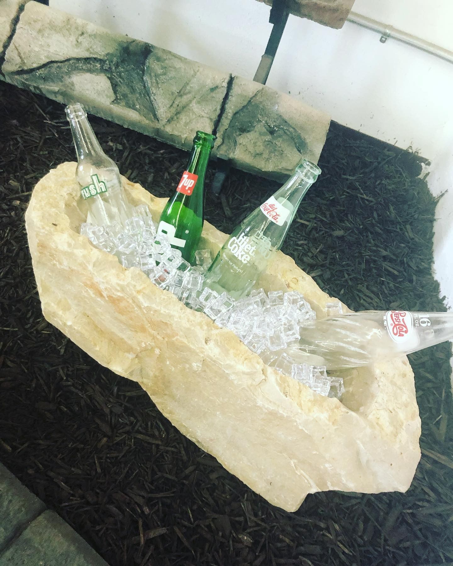 Glass soda bottles nestled in ice inside a carved stone basin resting on a bed of dark mulch.