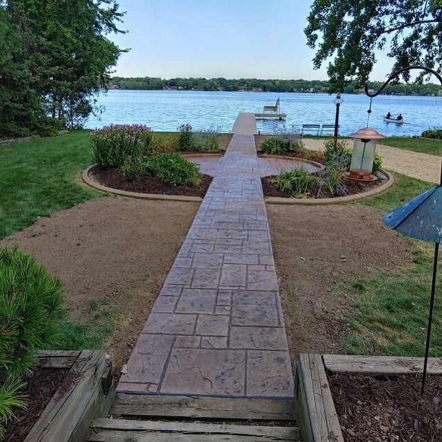 A stone pathway leads through a landscaped yard toward a wooden pier extending into a lake under a clear blue sky.