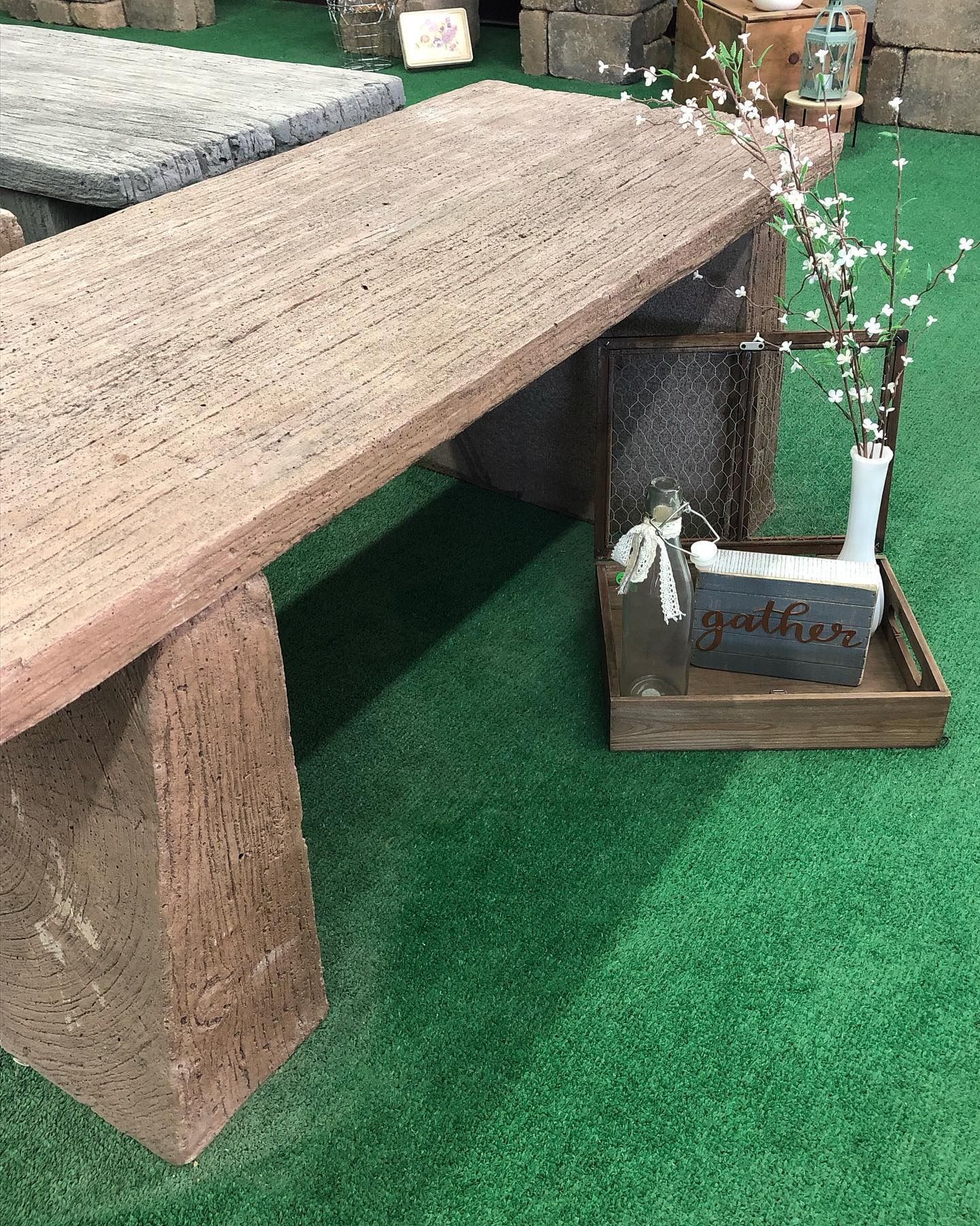 A rustic, textured stone bench sits on a green surface next to a decorative wooden crate holding a vase and sign.
