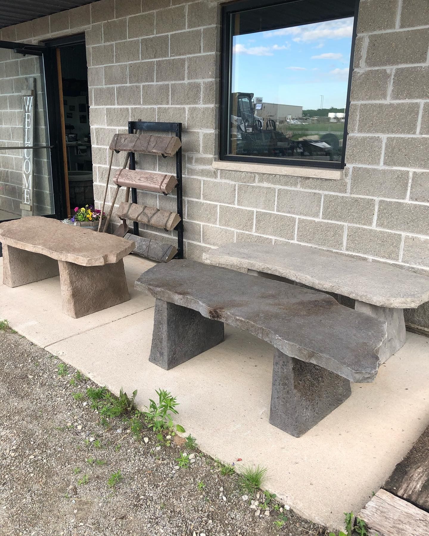 Two stone benches and a metal rack displaying stone slabs outside a building with a cinder block wall and a large window.